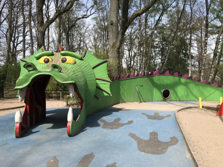 En lekplats med en stor drake i trä som är målad grön med lila taggar på ryggen. Barn kan gå in genom den öppna munnen med stora vita tänder. Marken framför är täckt av blått gummigolv med mörka fotavtryck, och runtom syns sand och höga träd i bakgrunden.